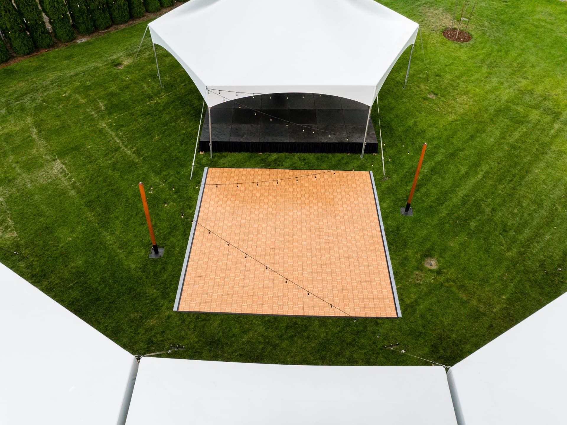 Aerial view of a black modular stage deck under a white high-peak tent with a parquet dance floor and bistro string lights at an Eastern Washington event
